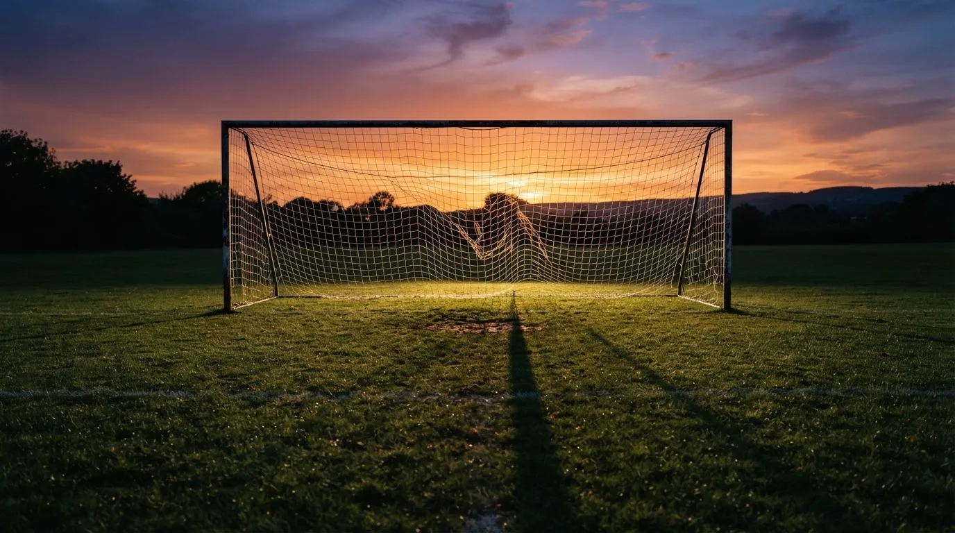 Ein Fußballtor mit Netz bei Sonnenuntergang auf einem leeren Platz, die Schatten der Torpfosten liegen lang auf dem Gras.
