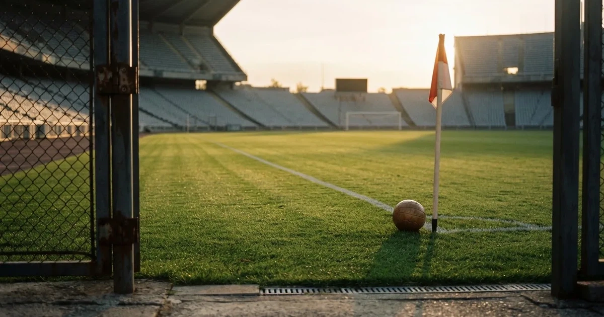 Blick durch ein Stadiontor auf einen grünen Rasen im Abendlicht, ein Ball ruht an der Eckfahne, die Tribünen sind leer.