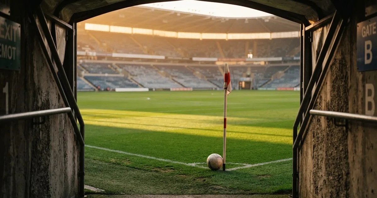 Blick durch ein Stadiontor auf einen grünen Rasen im Abendlicht, ein Ball liegt an der Eckfahne, die Ränge sind leer.