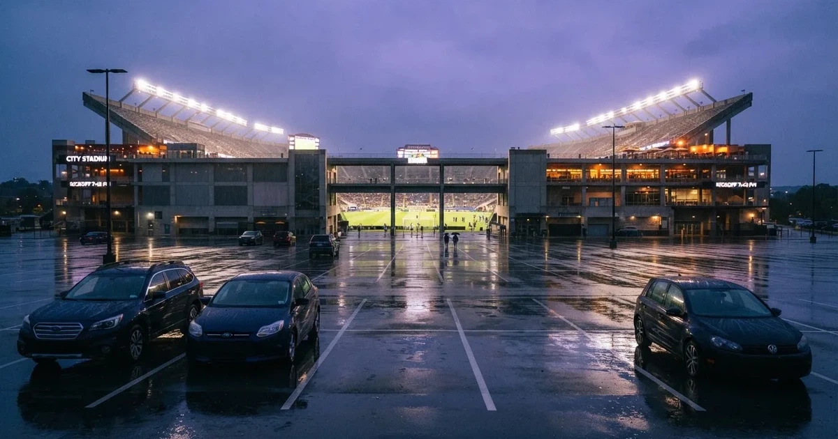 Ein Stadion unter Flutlicht aus großer Entfernung gesehen, davor ein regennasser Parkplatz in der Dämmerung.