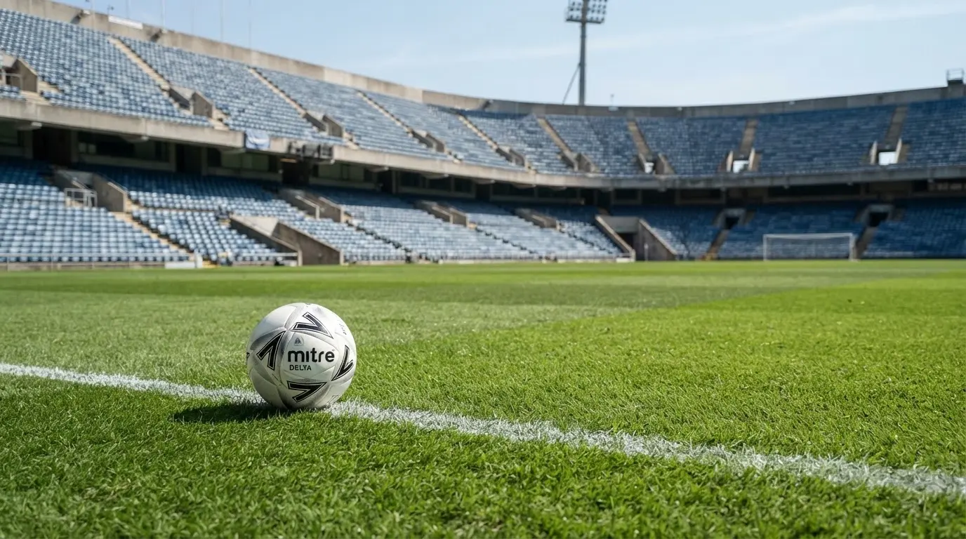 Ein Fußball liegt am Spielfeldrand eines Rasenplatzes, im Hintergrund eine leere Tribüne unter hellem Himmel.