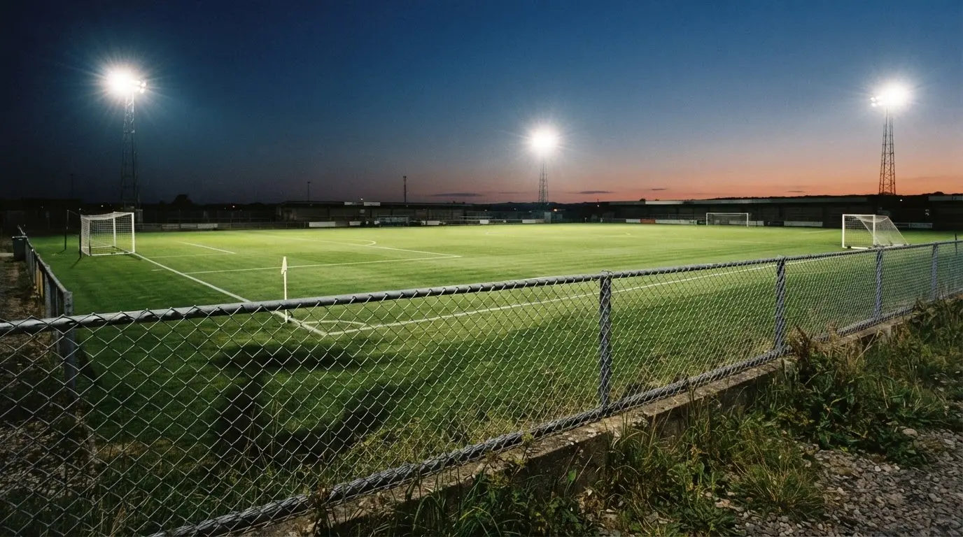 Eine Flutlichtanlage über einem leeren Sportplatz am Abend, im Vordergrund ein Maschendrahtzaun.