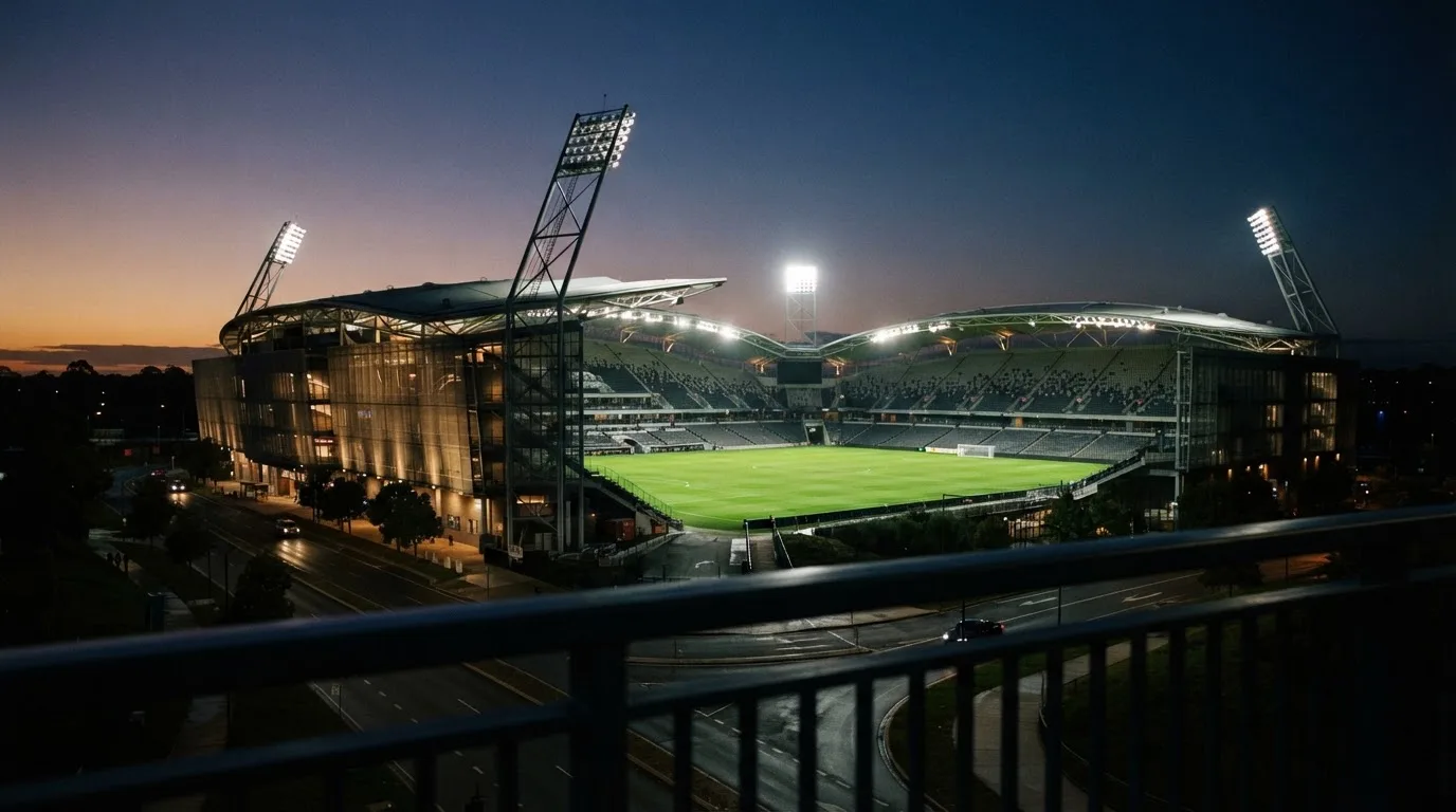 Ein Stadion bei Dämmerung mit eingeschalteten Flutlichtmasten, von einer Brücke aus aufgenommen.
