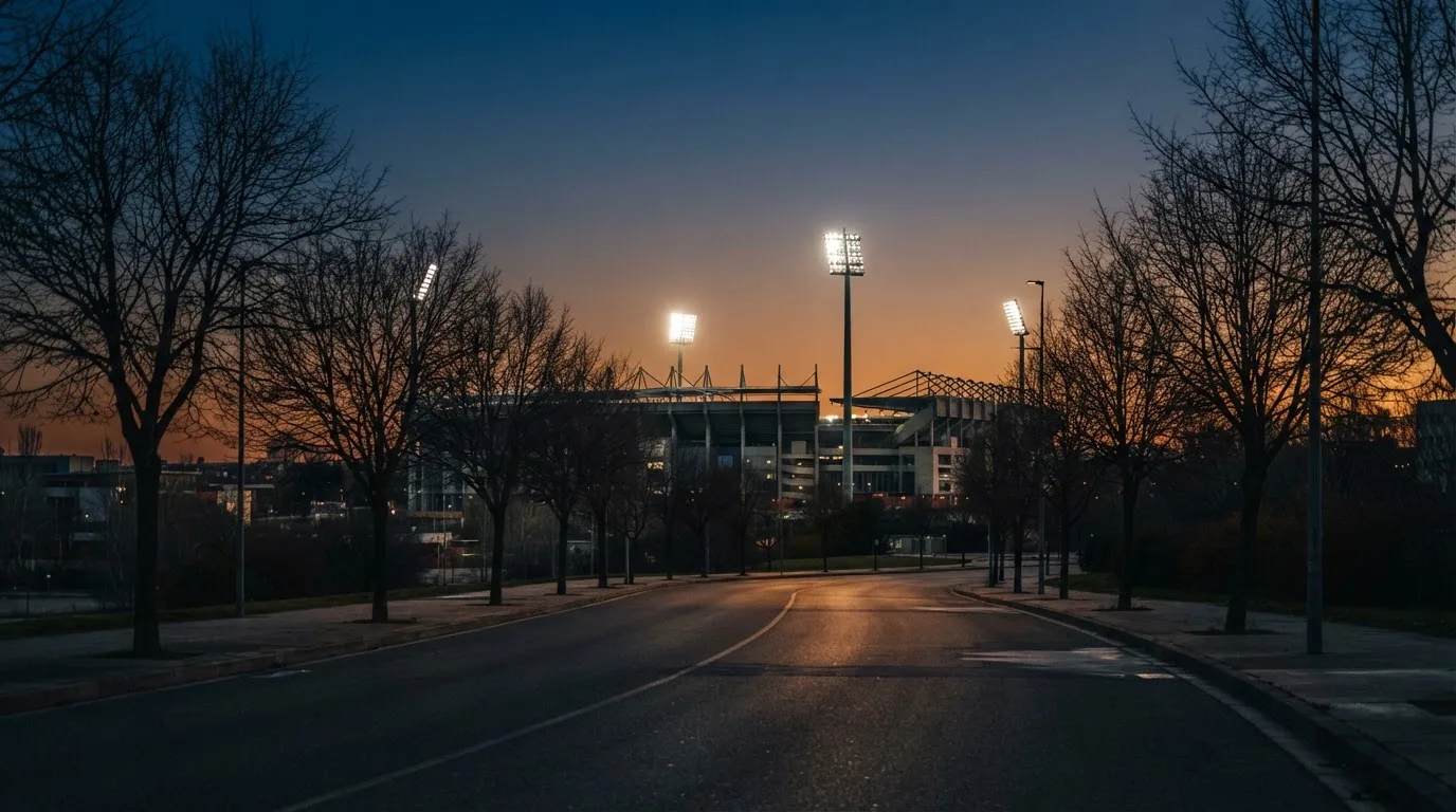Ein Stadion aus der Ferne bei Flutlicht, davor eine ruhige Straße in der Abenddämmerung.