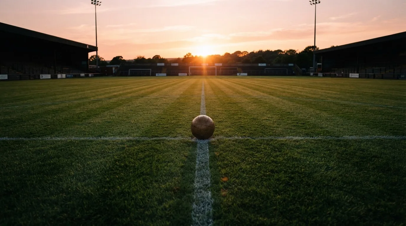 Ein Fußball liegt in der Mitte eines leeren Rasenplatzes, am Horizont färbt sich der Himmel orange.