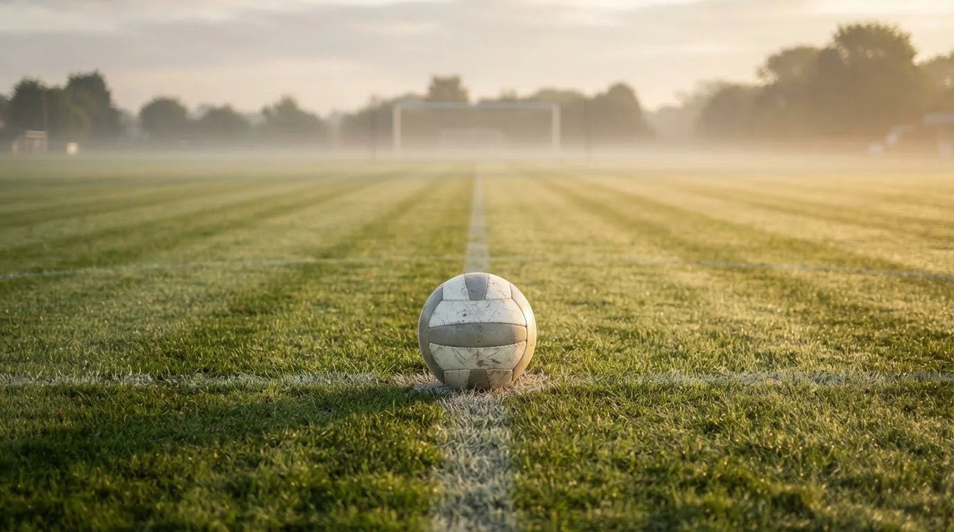 Ein Fußball auf dem Mittelkreis eines leeren Rasenplatzes im frühen Morgenlicht.