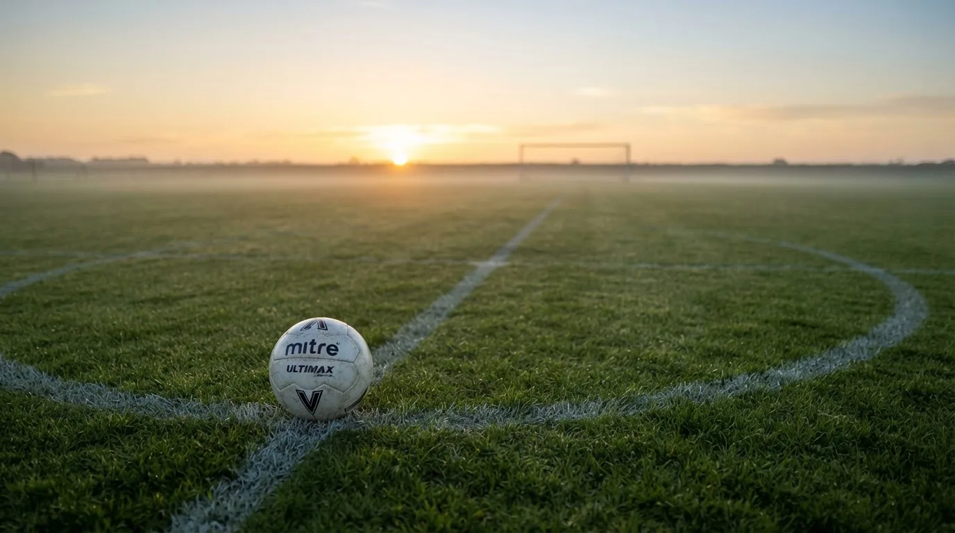 Ein Fußball liegt am Rand des Mittelkreises auf einem leeren Rasenplatz bei Sonnenaufgang.