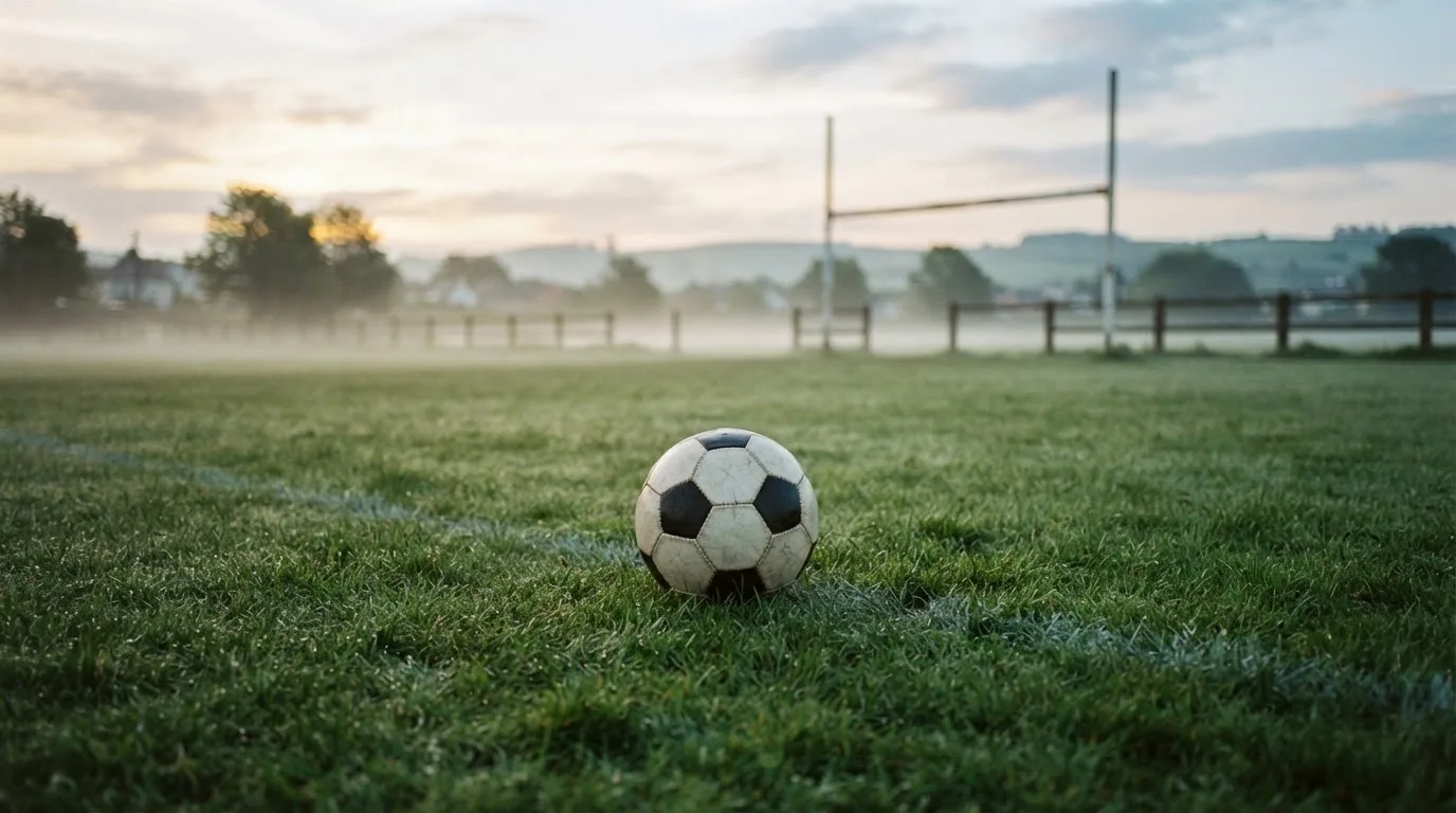 Ein Fußball auf dem Rasen eines leeren Sportplatzes am Rand einer Kleinstadt im Morgenlicht.
