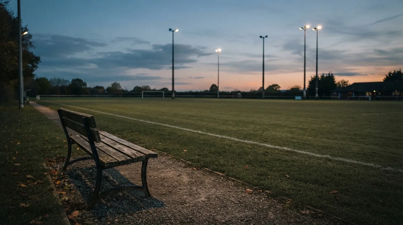 Eine leere Parkbank am Rand eines Fußballfelds in der Abenddämmerung, im Hintergrund ragen dunkle Flutlichtmasten in den Himmel.