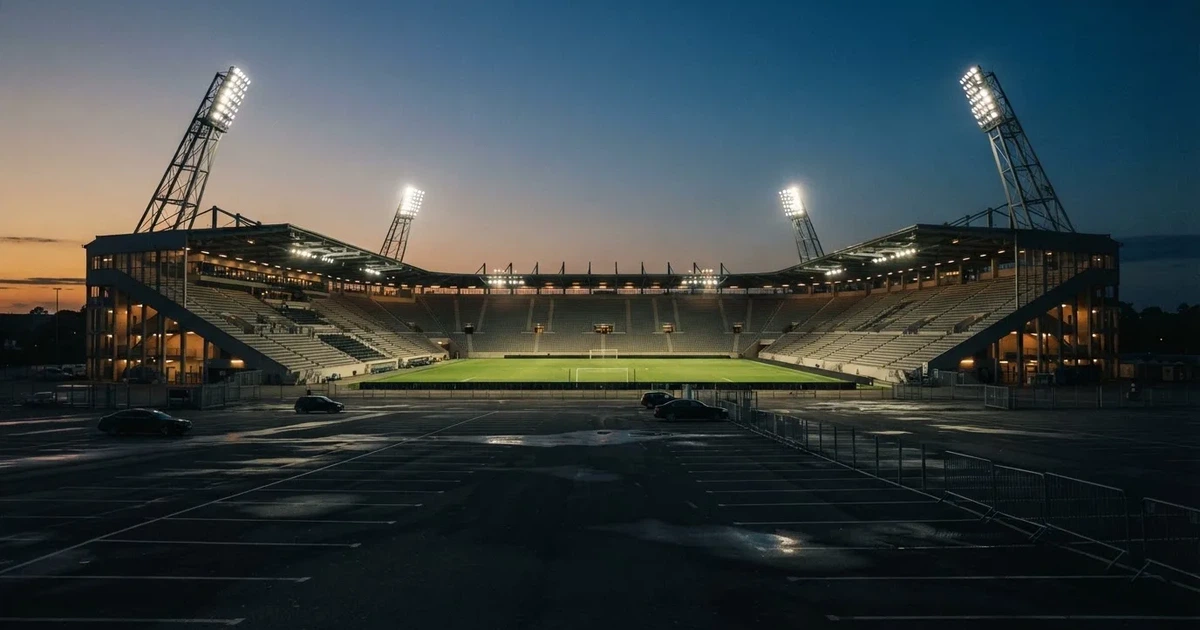 Ein Fußballstadion unter Flutlicht aus großer Entfernung, davor ein dunkler Parkplatz und ein heller Abendhimmel.