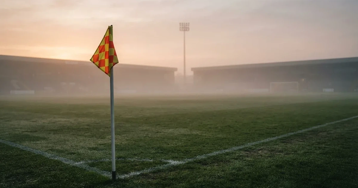 Eine Eckfahne steht im Nebel am Rand eines leeren Rasenplatzes in der Morgendämmerung, dahinter ein Flutlichtmast.