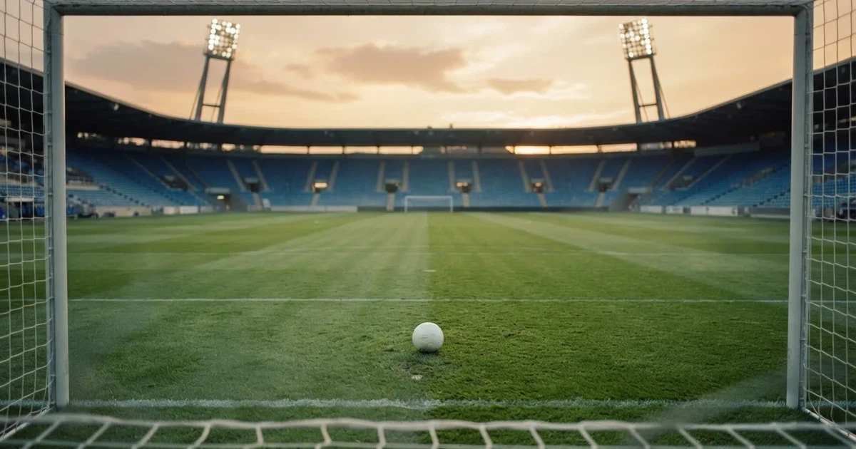Ein Fußballplatz mit frisch gemähtem Rasen im Abendlicht, ein Ball liegt im Strafraum, im Hintergrund unscharf eine leere Tribüne.