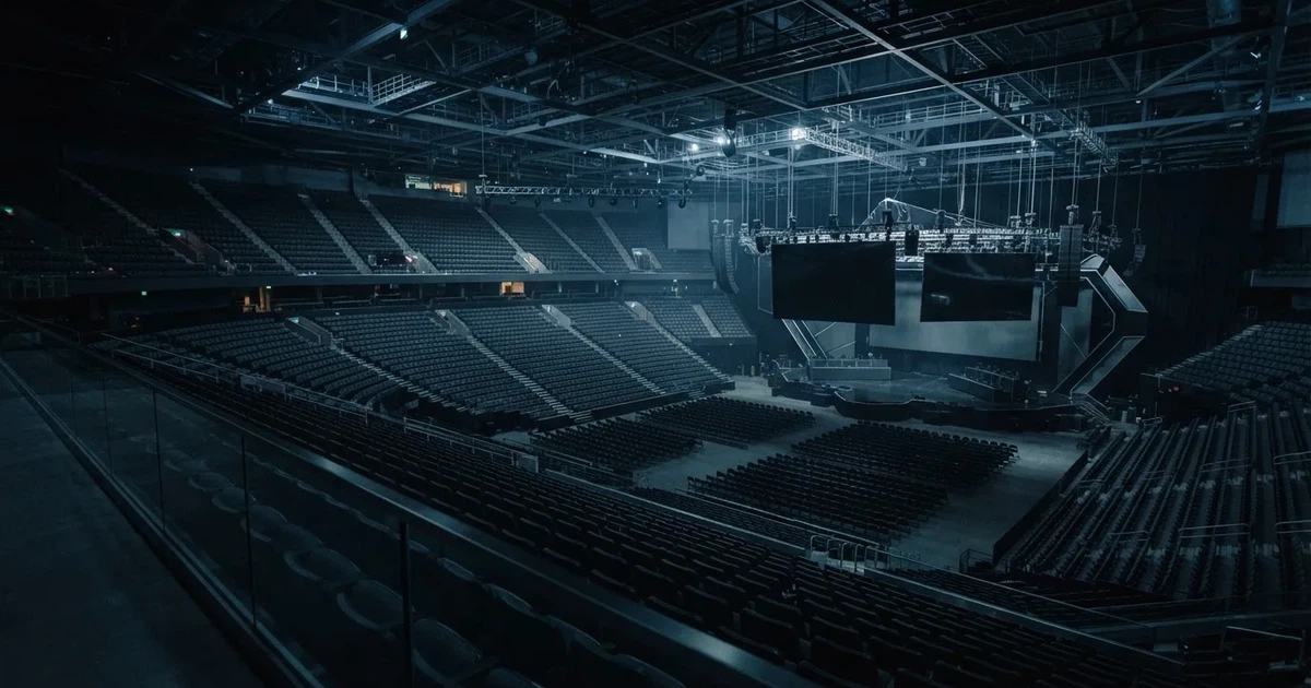 Eine große leere Arena mit Bühne und Stuhlreihen im Halbdunkel, von einem oberen Rang aus gesehen, alle Bildschirme ausgeschaltet.