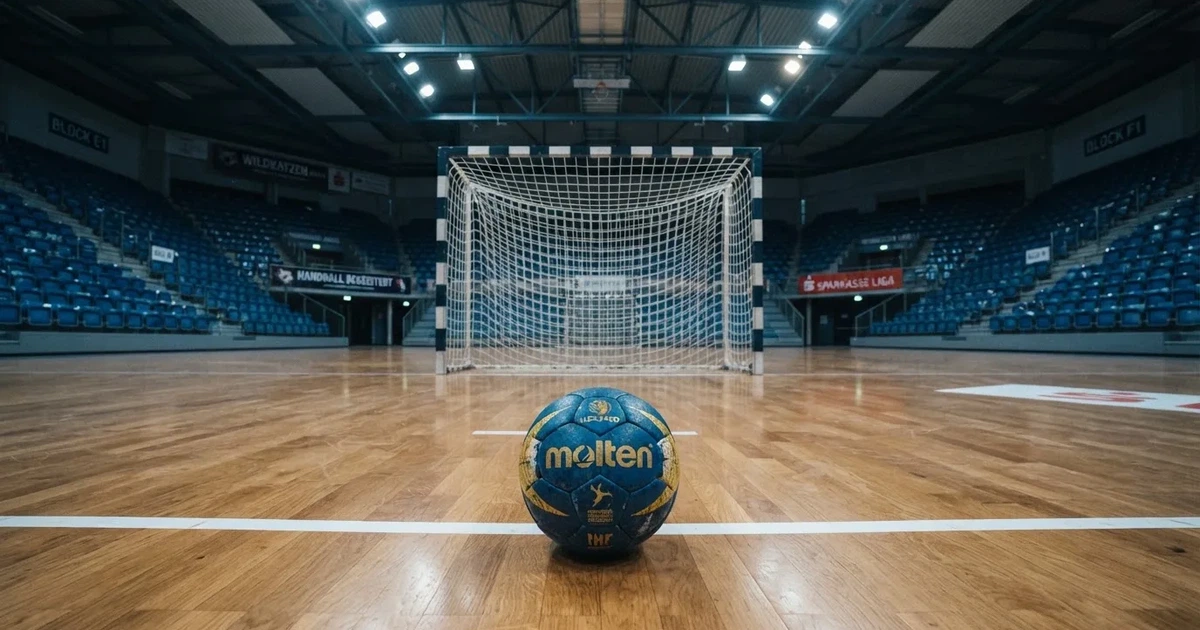 Ein Handball liegt auf dem Hallenboden vor einem Tor mit Netz, die leere Halle wird von Flutlicht beleuchtet.