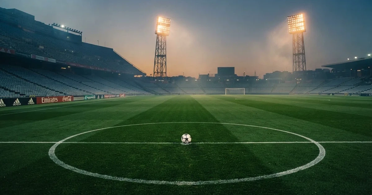 Ein Rasenplatz in der Dämmerung mit eingeschalteten Flutlichtmasten, ein Ball liegt am Anstoßpunkt, die Tribünen im Hintergrund sind unscharf.