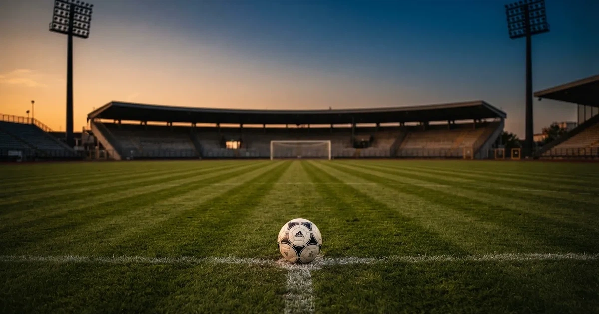 Ein Fußball liegt auf dem Anstoßpunkt eines leeren Rasenplatzes, im Hintergrund eine Tribüne und Flutlichtmasten in der Abenddämmerung.