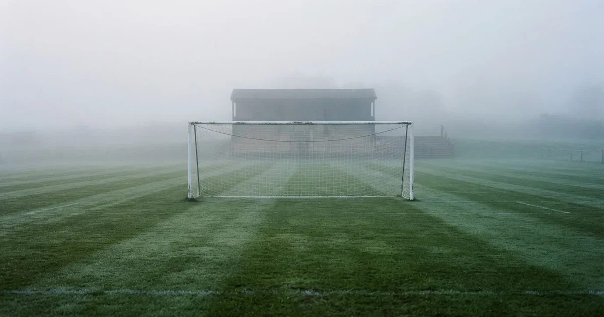 Ein Fußballtor mit Netz steht im Morgennebel auf einem leeren Rasenplatz, im Hintergrund die Silhouette einer kleinen Tribüne.