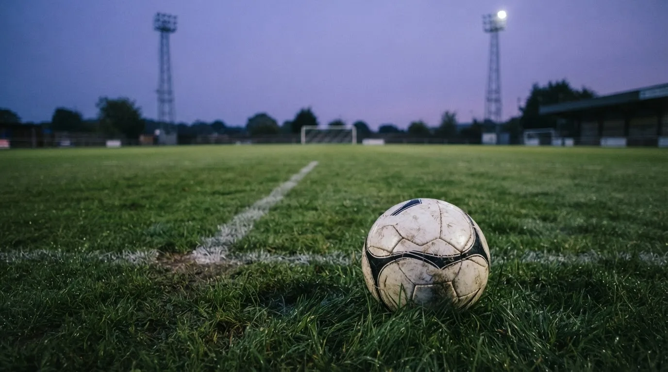 Ein Fußball liegt im Gras eines leeren Platzes, im Hintergrund stehen Flutlichtmasten in der Dämmerung.