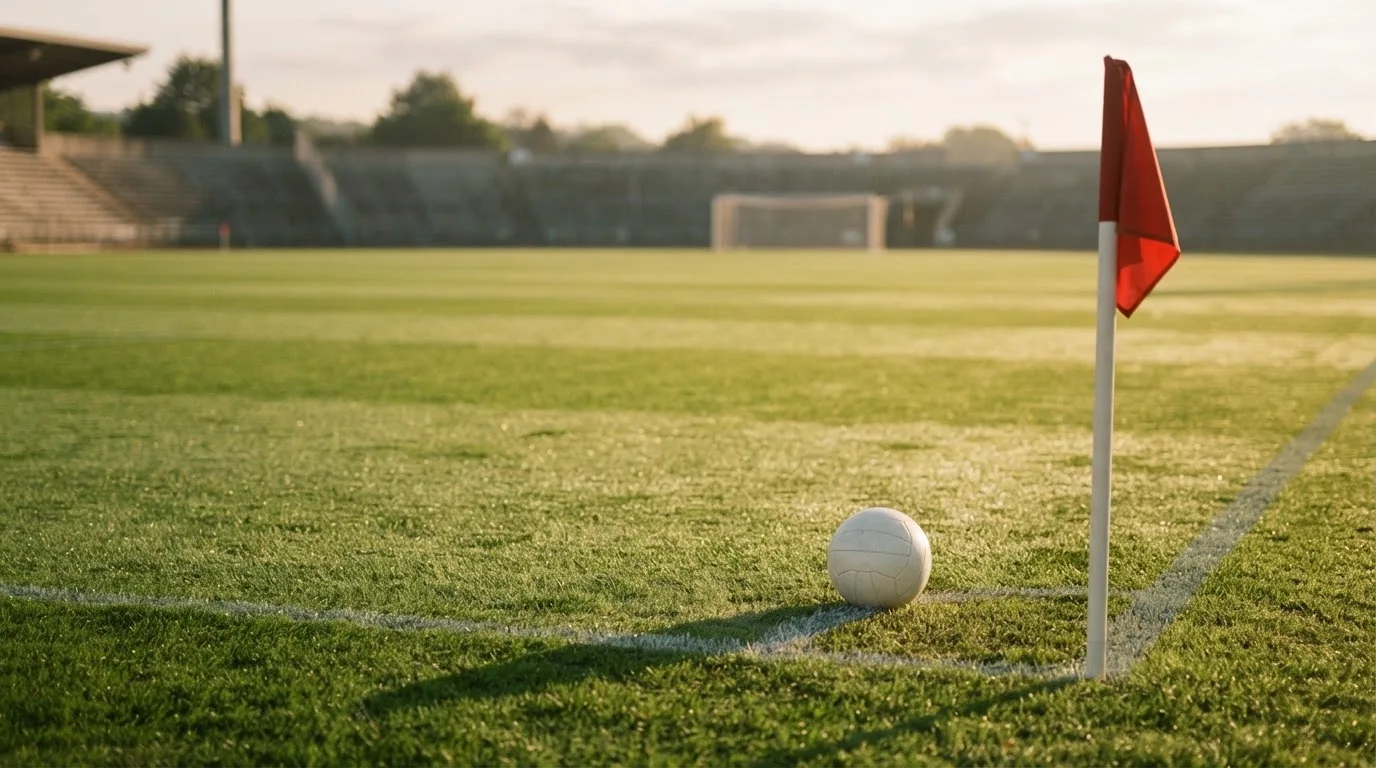 Ein Fußball liegt auf Rasen neben einer Eckfahne ohne Aufdruck auf einem leeren Platz im Morgenlicht.