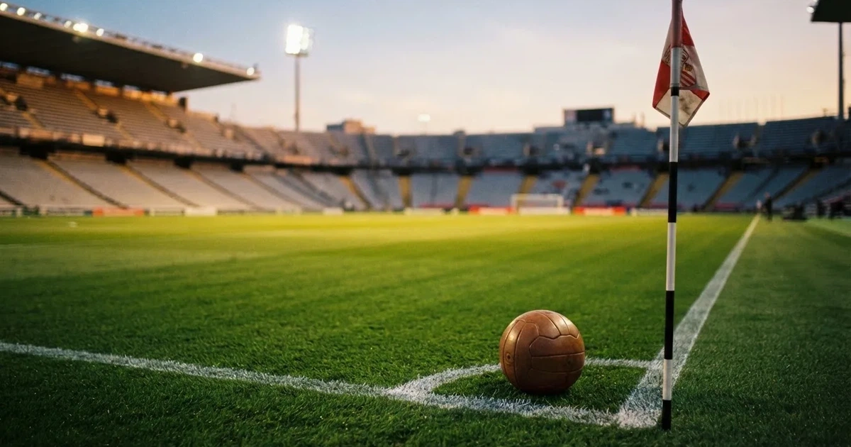 Ein Fußball liegt am Viertelkreis an der Eckfahne eines Rasenplatzes, im Hintergrund leere Tribünen in warmem Abendlicht.