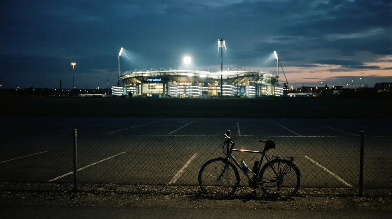 Ein Stadion unter Flutlicht aus der Ferne gesehen, davor ein dunkler Parkplatz und ein einzelnes Fahrrad an einem Zaun.