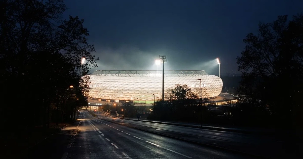 Ein Fußballstadion bei Nacht aus großer Entfernung, hell erleuchtet unter Flutlicht, davor eine dunkle Straße und Bäume.