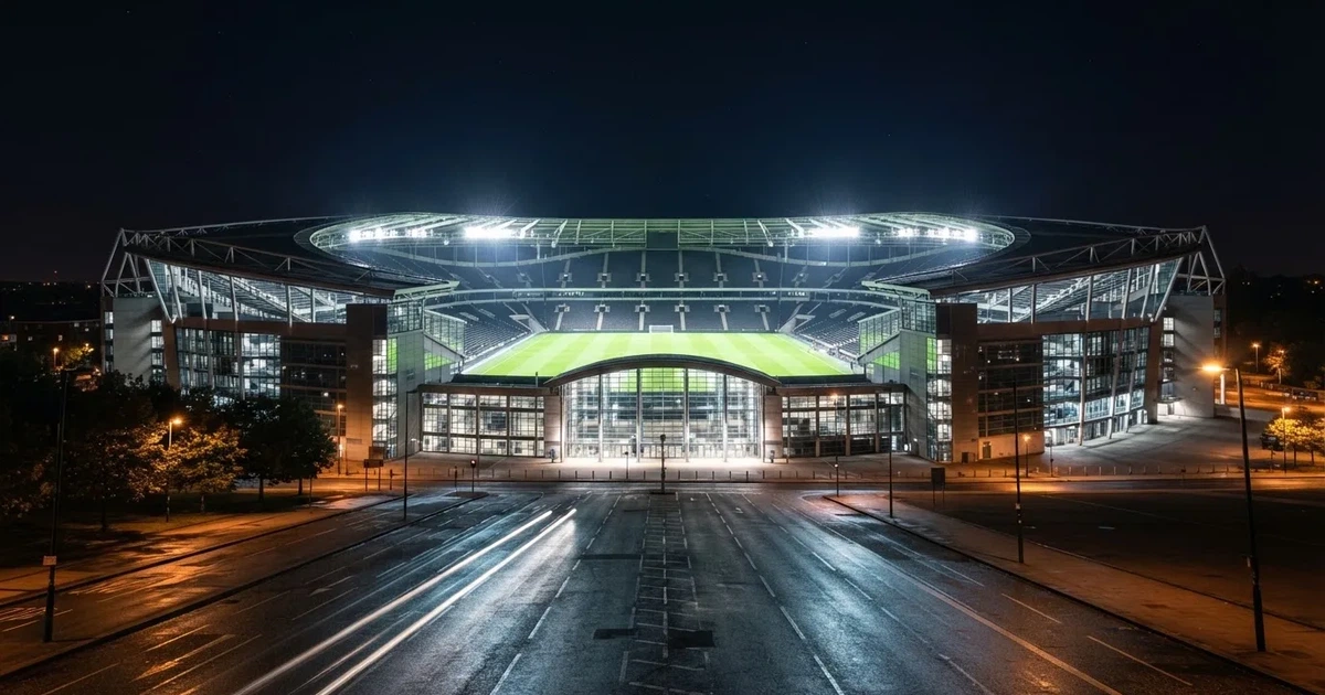 Ein hell erleuchtetes Fußballstadion bei Nacht aus der Ferne gesehen, über dem Dach ein klarer Himmel, davor eine leere Straße.