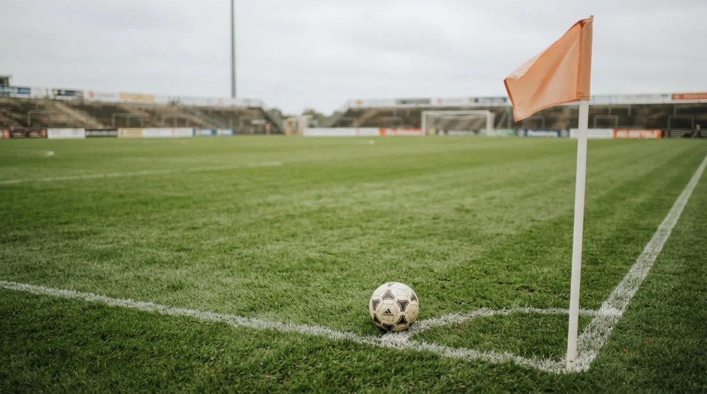 Ein Fußball liegt neben einer Eckfahne auf kurz gemähtem Rasen unter bewölktem Himmel.