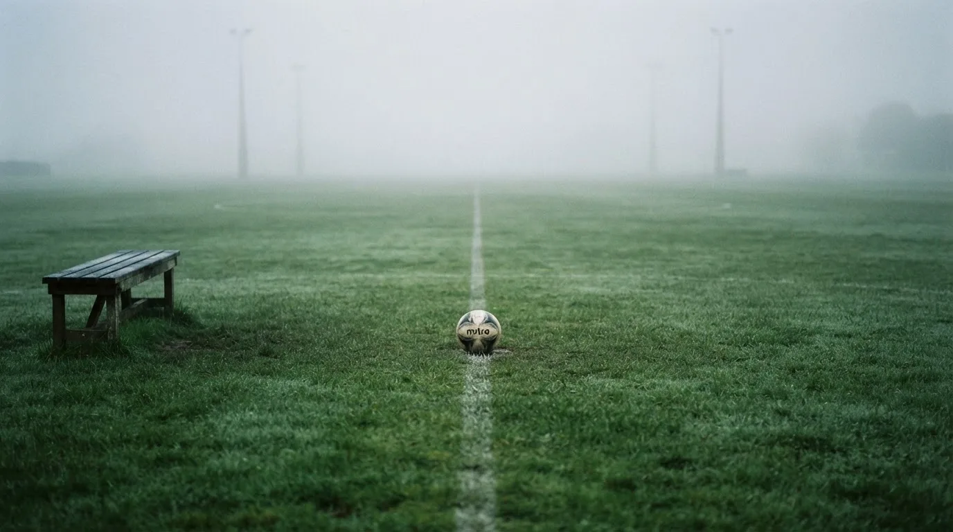 Ein Fußball liegt in der Mitte eines leeren Rasenplatzes im Morgennebel, am Rand steht eine einzelne Auswechselbank.